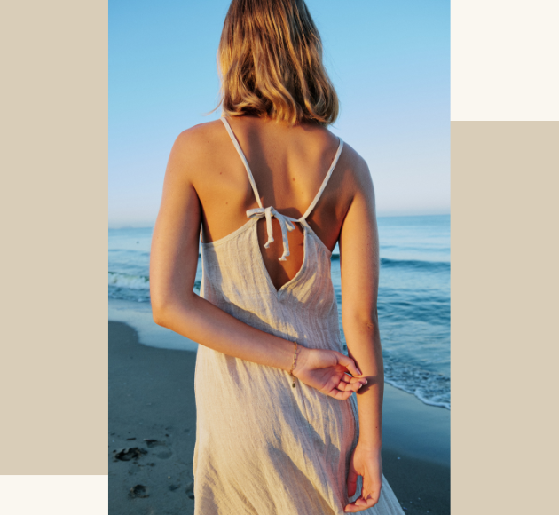 Modella fotografata di spalle con vestito di lino beige, mare sullo sfondo, atmosfera estiva e naturale