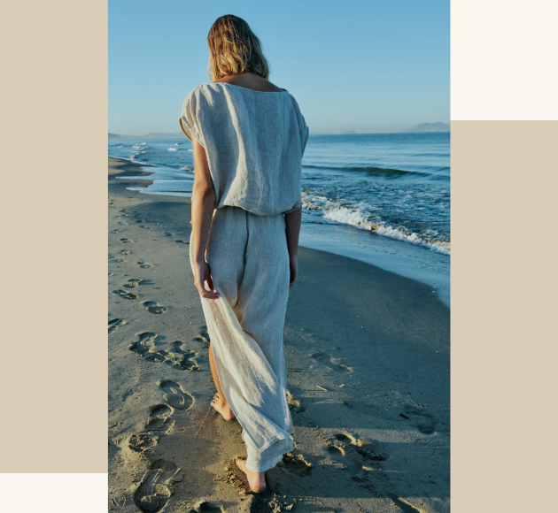 Modella fotografata di spalle che cammina sulla spiaggia con maglia a giro maniche beige e pantaloni di lino, atmosfera estiva e naturale
